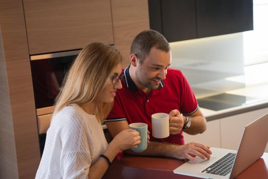 Young couple drinking coffee and using laptop computer at luxury home together, looking at screen, smiling.