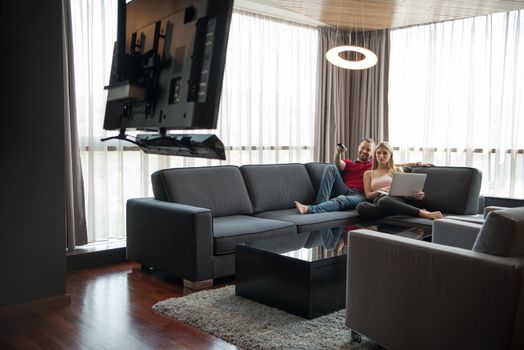 Young couple relaxes on the sofa in the luxury living room, using a laptop and remote control