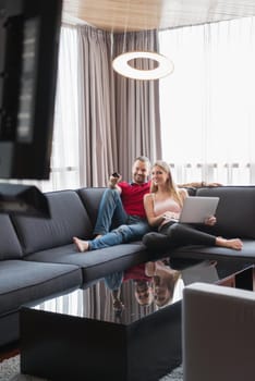 Young couple relaxes on the sofa in the luxury living room, using a laptop and remote control