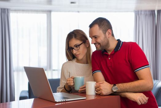 Young couple drinking coffee and using laptop computer at luxury home together, looking at screen, smiling.