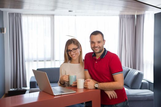 Young couple drinking coffee and using laptop computer at luxury home together, looking at screen, smiling.