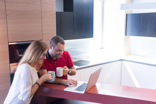 Young couple drinking coffee and using laptop computer at luxury home together, looking at screen, smiling.