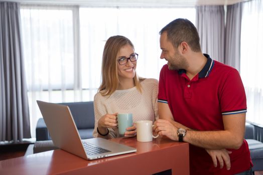 Young couple drinking coffee and using laptop computer at luxury home together, looking at screen, smiling.