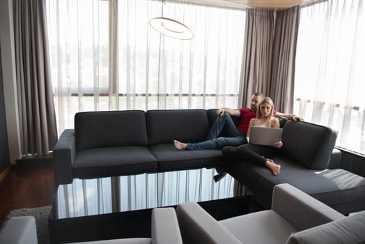 Young couple relaxes on the sofa in the luxury living room, using a laptop and remote control