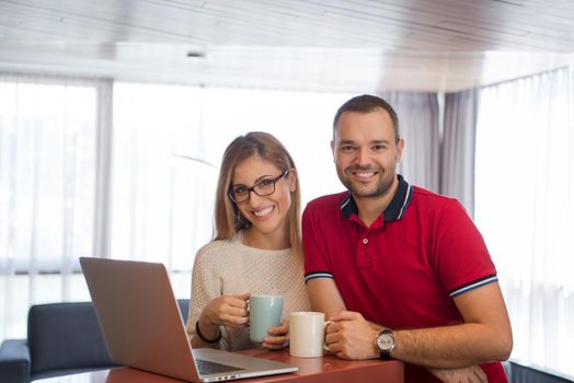 Young couple drinking coffee and using laptop computer at luxury home together, looking at screen, smiling.