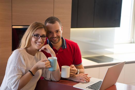 Young couple drinking coffee and using laptop computer at luxury home together, looking at screen, smiling.