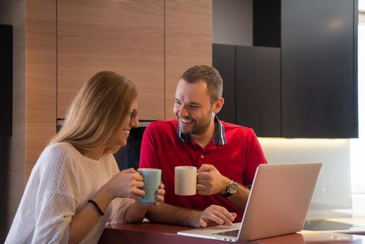 Young couple drinking coffee and using laptop computer at luxury home together, looking at screen, smiling.