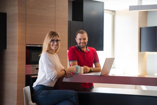 Young couple drinking coffee and using laptop computer at luxury home together, looking at screen, smiling.
