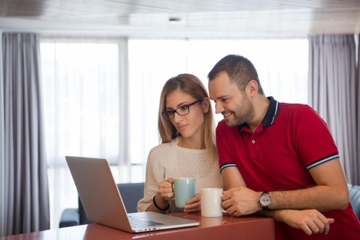 Young couple drinking coffee and using laptop computer at luxury home together, looking at screen, smiling.