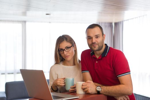 Young couple drinking coffee and using laptop computer at luxury home together, looking at screen, smiling.