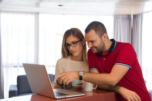 Young couple drinking coffee and using laptop computer at luxury home together, looking at screen, smiling.