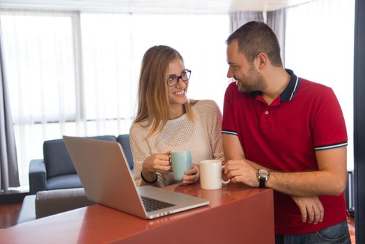 Young couple drinking coffee and using laptop computer at luxury home together, looking at screen, smiling.