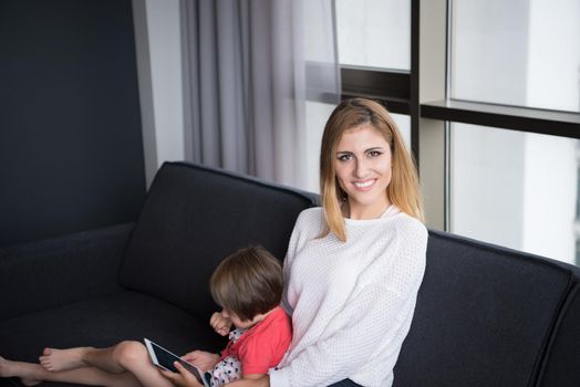 Beautiful young mother and her cute little daughter are using a tablet and smiling, sitting on sofa at home