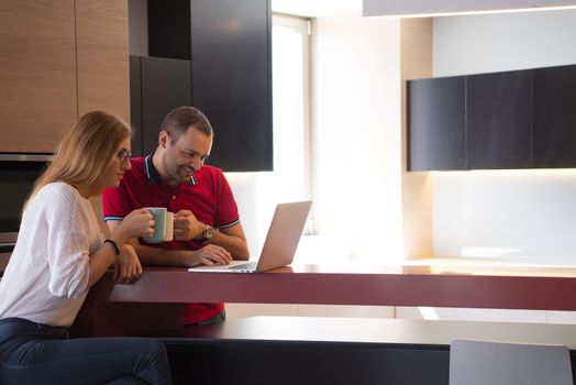 Young couple drinking coffee and using laptop computer at luxury home together, looking at screen, smiling.