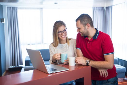 Young couple drinking coffee and using laptop computer at luxury home together, looking at screen, smiling.