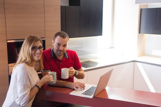 Young couple drinking coffee and using laptop computer at luxury home together, looking at screen, smiling.