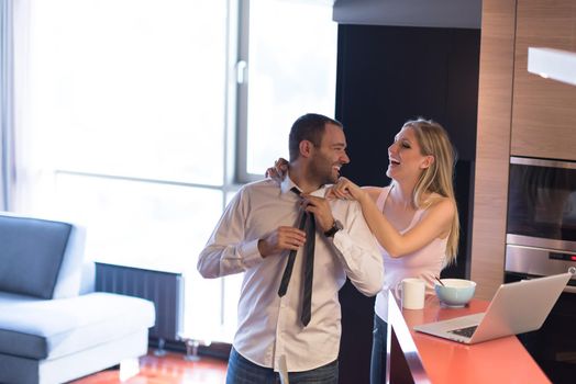 A young couple is preparing for the job and using a laptop. The man drinks coffee while the woman eats breakfast at luxury home together, looking at screen, smiling.