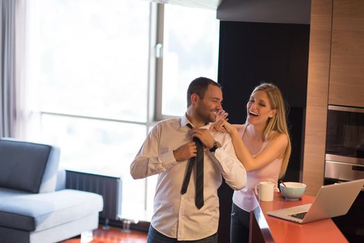 A young couple is preparing for the job and using a laptop. The man drinks coffee while the woman eats breakfast at luxury home together, looking at screen, smiling.