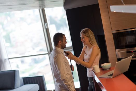 A young couple is preparing for the job and using a laptop. The man drinks coffee while the woman eats breakfast at luxury home together, looking at screen, smiling.