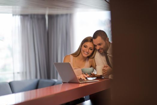 A young couple is preparing for the job and using a laptop. The man drinks coffee while the woman eats breakfast at luxury home together, looking at screen, smiling.