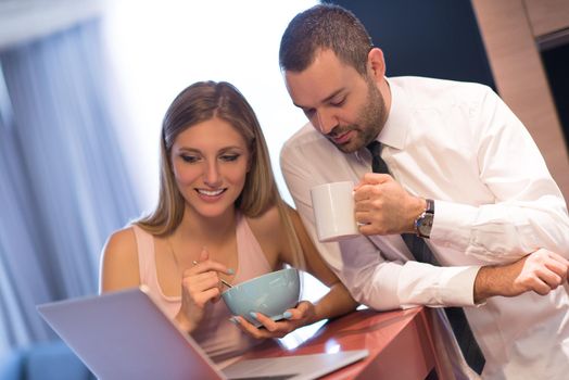 A young couple is preparing for the job and using a laptop. The man drinks coffee while the woman eats breakfast at luxury home together, looking at screen, smiling.