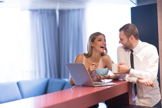 A young couple is preparing for the job and using a laptop. The man drinks coffee while the woman eats breakfast at luxury home together, looking at screen, smiling.