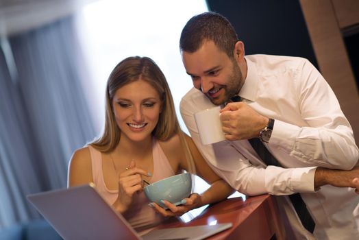 A young couple is preparing for the job and using a laptop. The man drinks coffee while the woman eats breakfast at luxury home together, looking at screen, smiling.