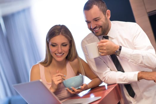 A young couple is preparing for the job and using a laptop. The man drinks coffee while the woman eats breakfast at luxury home together, looking at screen, smiling.