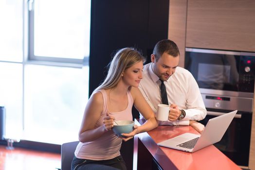 A young couple is preparing for the job and using a laptop. The man drinks coffee while the woman eats breakfast at luxury home together, looking at screen, smiling.