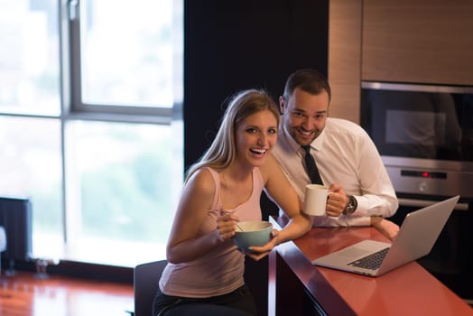 A young couple is preparing for the job and using a laptop. The man drinks coffee while the woman eats breakfast at luxury home together, looking at screen, smiling.