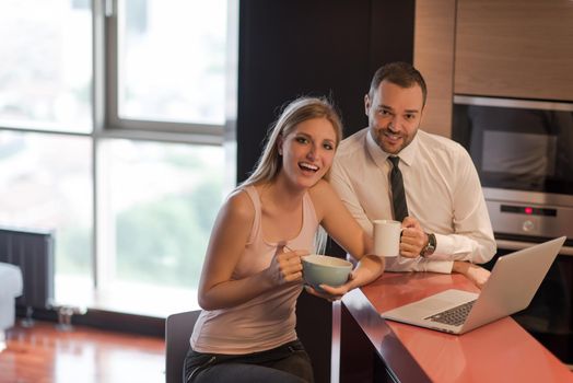 A young couple is preparing for the job and using a laptop. The man drinks coffee while the woman eats breakfast at luxury home together, looking at screen, smiling.