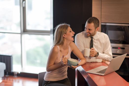 A young couple is preparing for the job and using a laptop. The man drinks coffee while the woman eats breakfast at luxury home together, looking at screen, smiling.