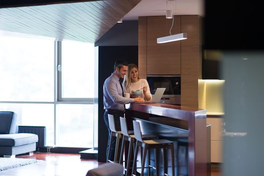 A young couple is preparing for the job and using a laptop. The man drinks coffee while the woman eats breakfast at luxury home together, looking at screen, smiling.