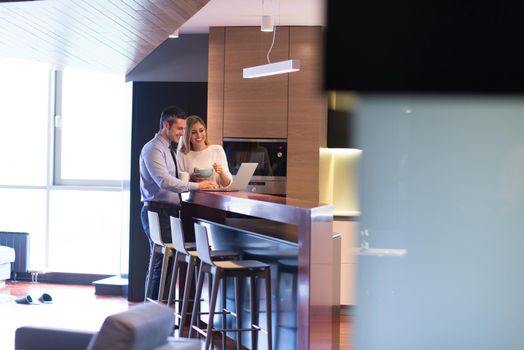 A young couple is preparing for the job and using a laptop. The man drinks coffee while the woman eats breakfast at luxury home together, looking at screen, smiling.