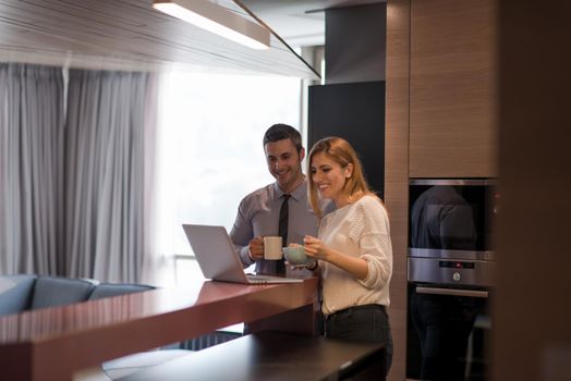 A young couple is preparing for the job and using a laptop. The man drinks coffee while the woman eats breakfast at luxury home together, looking at screen, smiling.