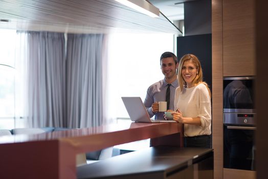 A young couple is preparing for the job and using a laptop. The man drinks coffee while the woman eats breakfast at luxury home together, looking at screen, smiling.