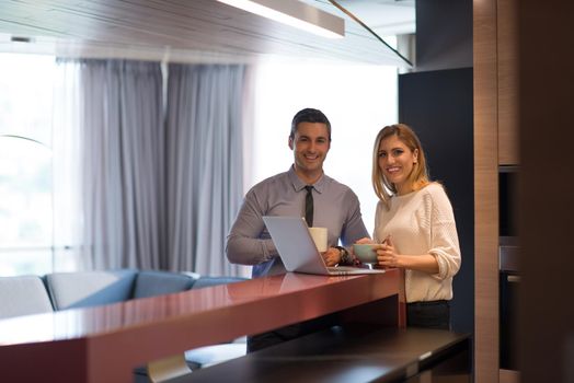 A young couple is preparing for the job and using a laptop. The man drinks coffee while the woman eats breakfast at luxury home together, looking at screen, smiling.