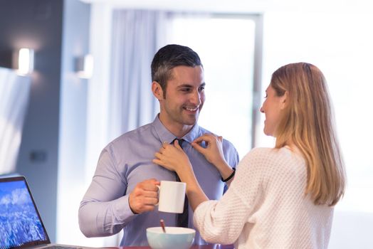 A young couple is preparing for the job and using a laptop. The man drinks coffee while the woman eats breakfast at luxury home together, looking at screen, smiling.