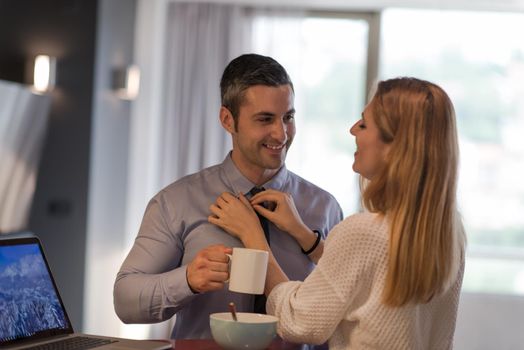 A young couple is preparing for the job and using a laptop. The man drinks coffee while the woman eats breakfast at luxury home together, looking at screen, smiling.