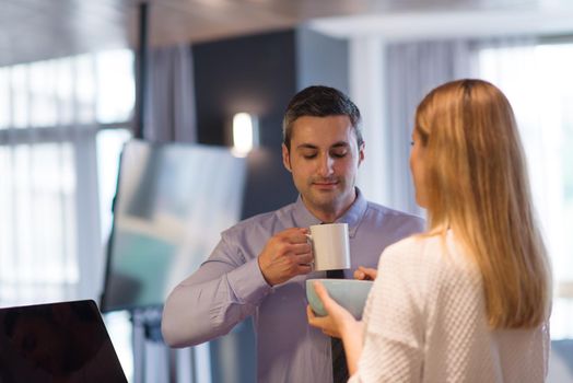 A young couple is preparing for the job and using a laptop. The man drinks coffee while the woman eats breakfast at luxury home together, looking at screen, smiling.