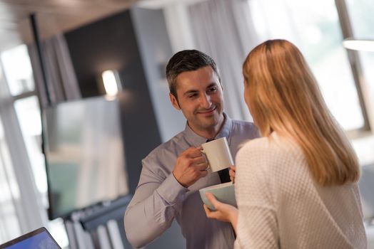 A young couple is preparing for the job and using a laptop. The man drinks coffee while the woman eats breakfast at luxury home together, looking at screen, smiling.