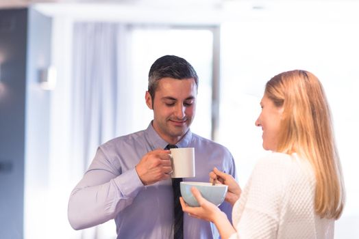 A young couple is preparing for the job and using a laptop. The man drinks coffee while the woman eats breakfast at luxury home together, looking at screen, smiling.