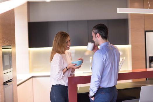 A young couple is preparing for the job and using a laptop. The man drinks coffee while the woman eats breakfast at luxury home together, looking at screen, smiling.