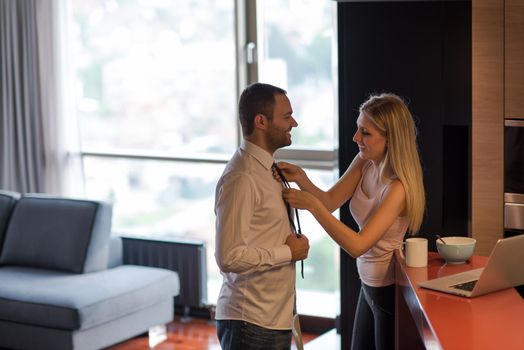 A young couple is preparing for the job and using a laptop. The man drinks coffee while the woman eats breakfast at luxury home together, looking at screen, smiling.