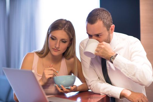 A young couple is preparing for the job and using a laptop. The man drinks coffee while the woman eats breakfast at luxury home together, looking at screen, smiling.