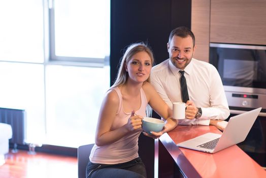 A young couple is preparing for the job and using a laptop. The man drinks coffee while the woman eats breakfast at luxury home together, looking at screen, smiling.