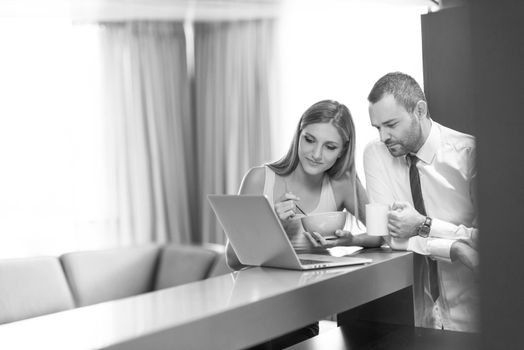 A young couple is preparing for the job and using a laptop. The man drinks coffee while the woman eats breakfast at luxury home together, looking at screen, smiling.