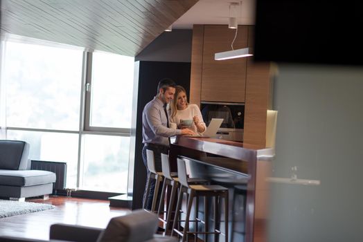 A young couple is preparing for the job and using a laptop. The man drinks coffee while the woman eats breakfast at luxury home together, looking at screen, smiling.