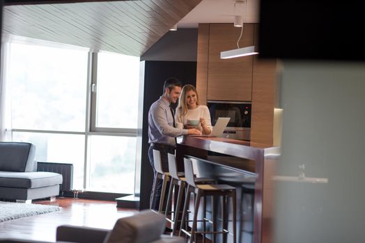 A young couple is preparing for the job and using a laptop. The man drinks coffee while the woman eats breakfast at luxury home together, looking at screen, smiling.