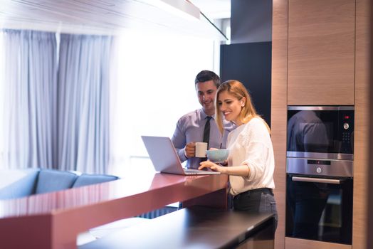 A young couple is preparing for the job and using a laptop. The man drinks coffee while the woman eats breakfast at luxury home together, looking at screen, smiling.